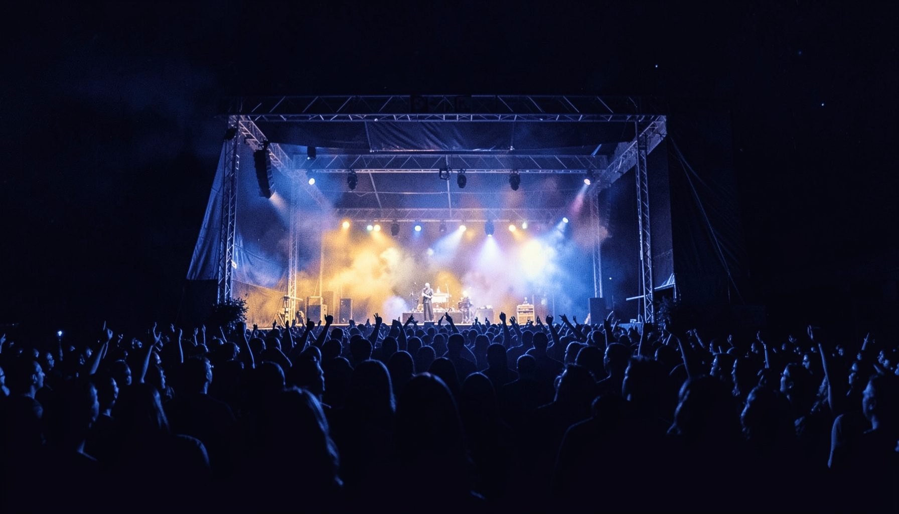 A dark, atmospheric duotone photograph of a festival stage lit in deep indigo and warm amber, with a crowd silhouetted against the glow.
