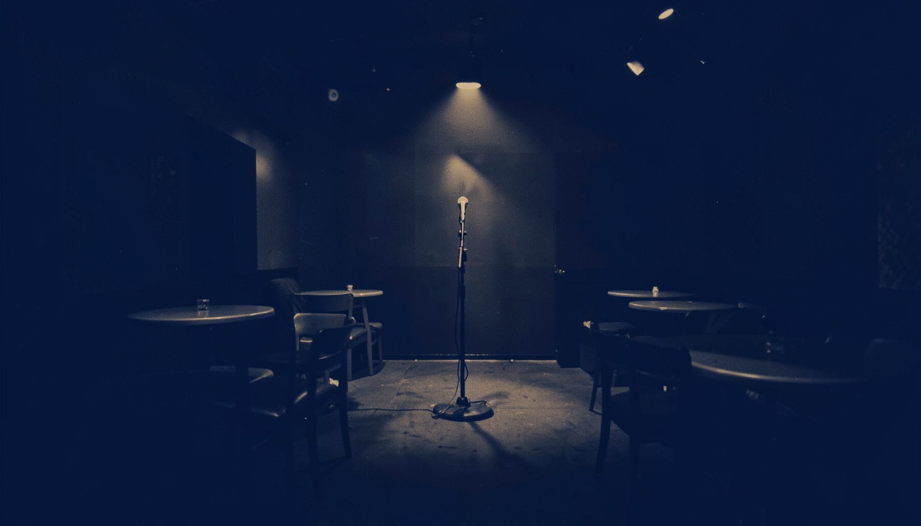A warm duotone photograph of a dimly lit jazz club interior, deep navy and muted amber tones, with empty round tables and a lone microphone stand on a small stage.
